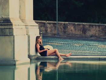Full length side view of woman sitting by pond in city