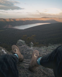 Low section of man sitting on mountain against sky during sunset