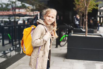 After school, a cute schoolgirl with a backpack stands near a glass building while waiting for a bus