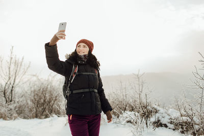 Smiling woman in warm taking selfie while standing on snowcapped mountain