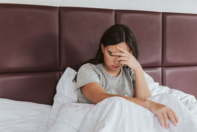 Depressed young woman sitting in bed, holding face in hands.