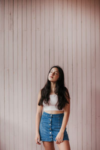 Calm dreamy female teenager with long dark hair wearing casual crop top and denim skirt standing with eyes closed against plank wall