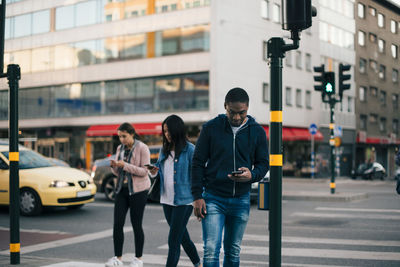 People crossing street while using smart phone against building in city