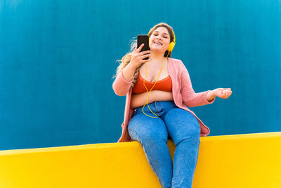 Young woman using mobile phone while sitting against wall