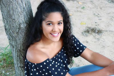 Portrait of young woman smiling while sitting by tree trunk at beach