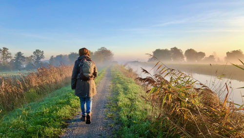 Rear view of a woman walking at sunrise over a green dike along a misty canal