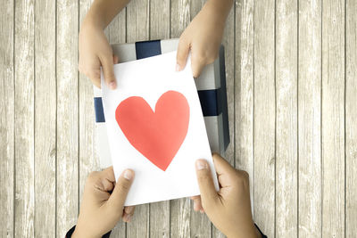 Cropped hands holding heart shape paper over gift on table