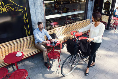 Businesswoman with bicycle talking to male colleague sitting at sidewalk cafe