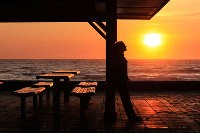 Silhouette woman leaning on pole at beach against sky during sunset