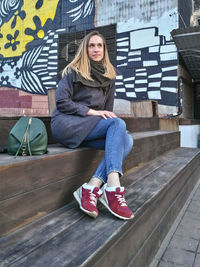 A young woman in a coat, jeans and sneakers sits on the wooden steps of a painted wall