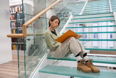 Focused female student enjoys reading interesting book with orange cover sitting on glass stairs