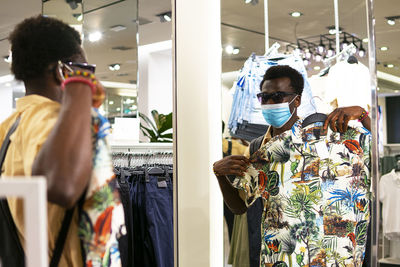 Young african american male in trendy outfit and medical mask standing in front of mirror in modern boutique and picking goods in shop during coronavirus