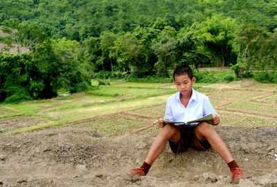 Full length of cute boy reading book while sitting on field against trees