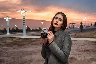 Portrait of young woman photographing against sky during sunset