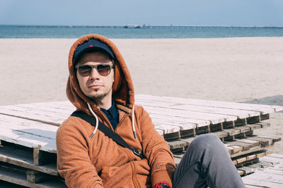 Portrait of young man sitting at beach against sky