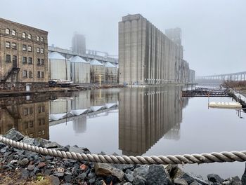 Industrial buildings in a shipyard reflected in water on an overcast day. 