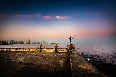 People fishing on pier by sea against sky during sunset