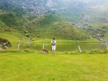 Rear view of man with dog standing on grassy field by lake and mountain