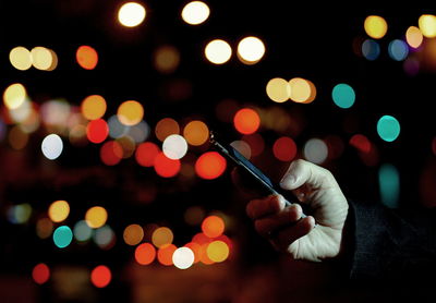 Cropped hand of man using mobile phone against illuminated lights at night