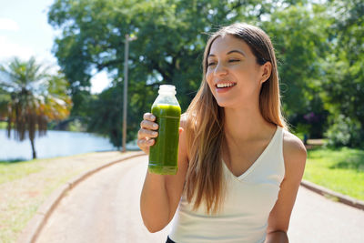 Portrait of healthy woman holding a bottle of green smoothie detox juice homemade outdoors. 