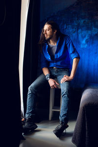 Man with long hair and beard sits at home on a chair by the window in a blue shirt and jeans