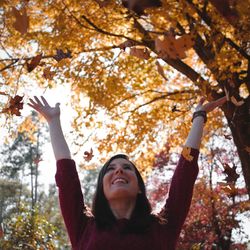Smiling young woman playing with maple leaves in park during autumn