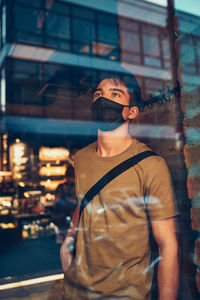 Young man standing in coffee shop at store front in the city center in the evening, wearing the mask