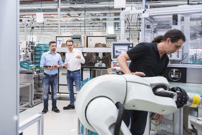 Man operating assembly robot in factory with two men in background supervising