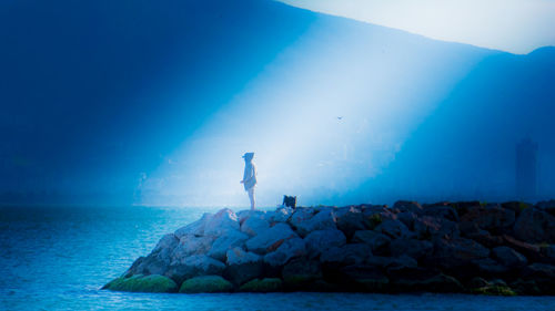 Man standing on rock by sea against blue sky