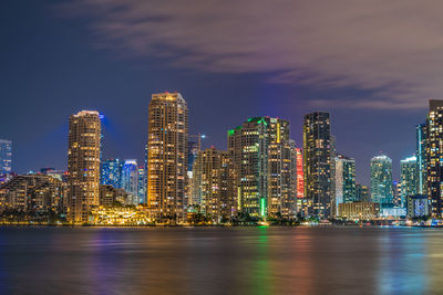 Illuminated modern buildings by sea against sky at night