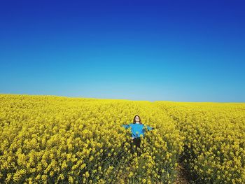 Young woman with arms outstretched standing by flowers on field against clear blue sky