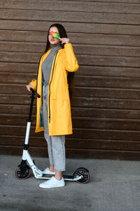 A girl in a yellow raincoat stands on a white scooter and holds colorful ice cream in her hands