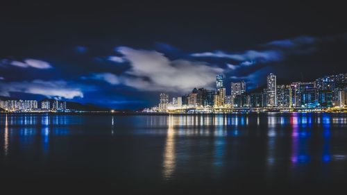 Illuminated modern buildings in front of sea against cloudy sky at night in city