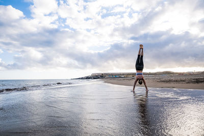 Full length of young woman doing handstand at beach against cloudy sky