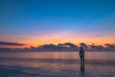 Rear view of man standing on shore at beach against sky during sunset