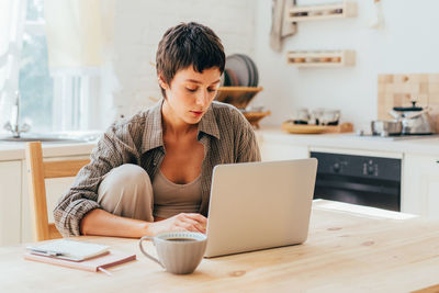 Young short-haired modern woman working on the computer while sitting at the table in the kitchen