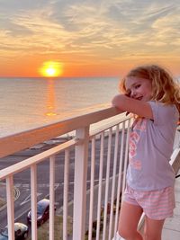 Portrait of young girl standing on railing against sea during sunset