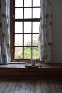 A coffee cup on a handmade tray on a window sill. paned wooden window with curtains, wooden floor.