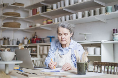 Senior female potter writing on paper while sitting with laptop at table in workshop