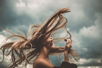 Side view of young woman with tousled hair standing against cloudy sky