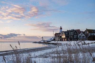 Houses by frozen lake and buildings against sky during sunset