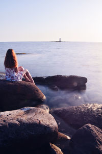 Rear view of woman sitting on rock by sea against sky