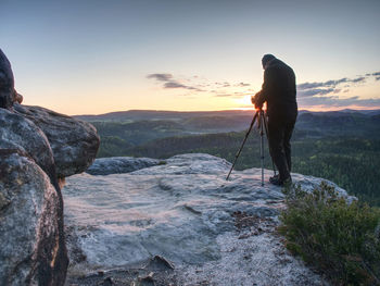 Man takes photos with camera on tripod on rocky peak tourist with camera and tripod in cold weather