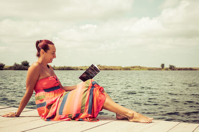 Smiling woman in sun dress reading book while sitting on a pier during summer day. copy space.