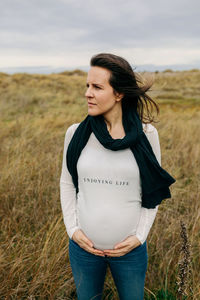 Pregnant young woman touching abdomen while standing on grass against sky