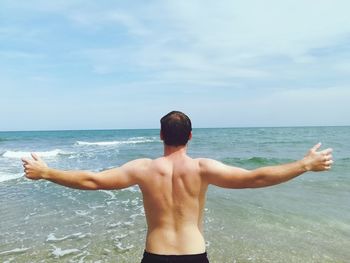 Rear view of shirtless man standing with arms outstretched by sea