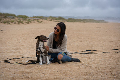 Caucasian woman with sunglasses sit and playing with a brown dog in the sand of lanzada beach