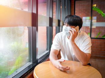A handsome young teenage boy wear mask talkinging on smart mobile phone waiting in cafeteria. 