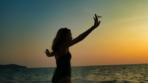 Side view of woman with arms raised standing at beach against sky during sunset