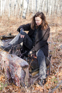 A young woman in casual attire sits on an old suitcase in a serene forest. 